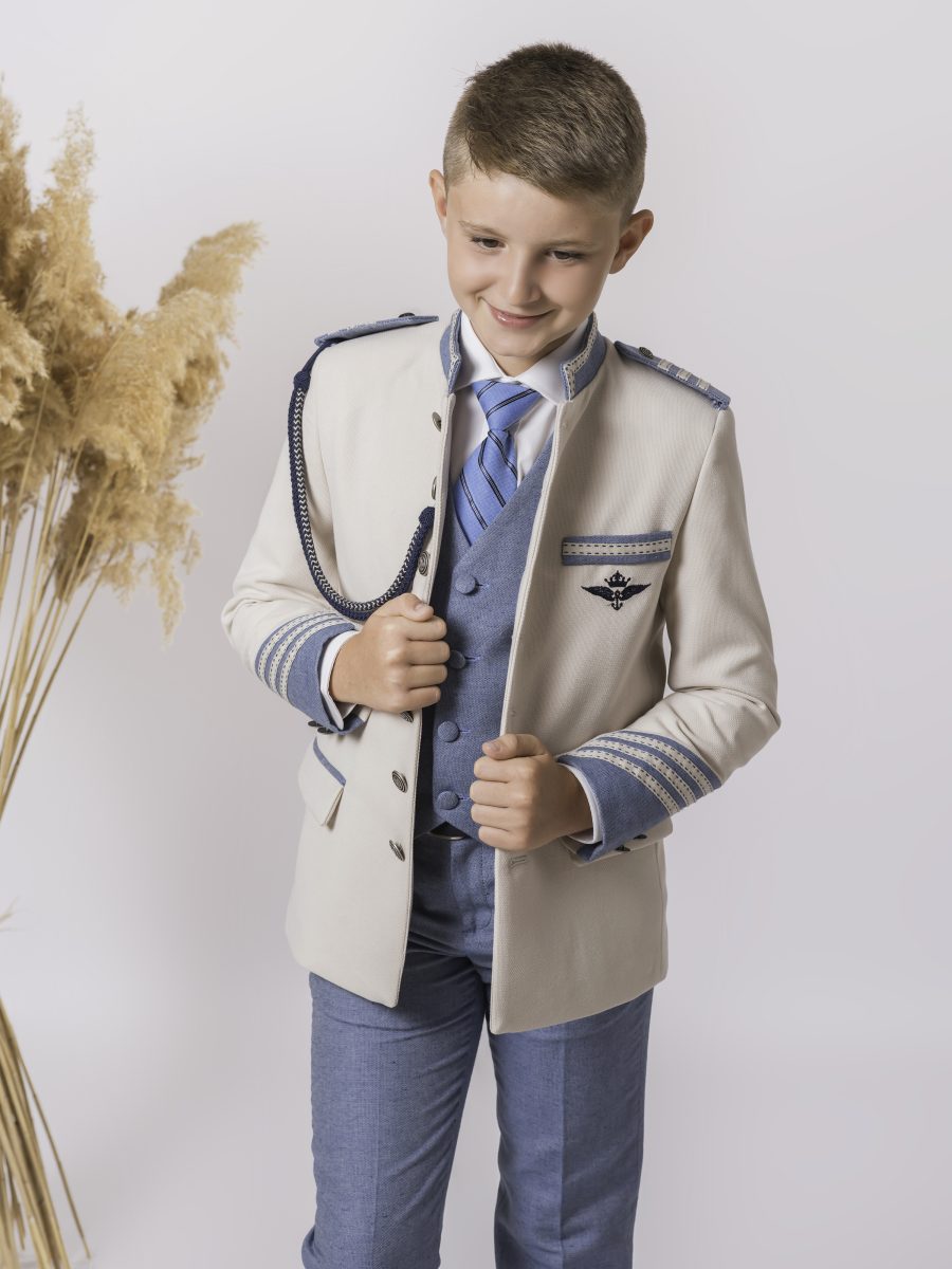 Chaqueta de comunión beige crema con ribetes azul celeste, cordón bicolor e insignia en el pecho, combinada con chaleco azul y beige.