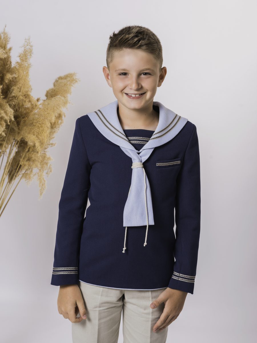 Detalle de blusa marinera azul marino de comunión con cuello y corbata azul celeste, ribetes decorativos y pantalón beige como contraste.