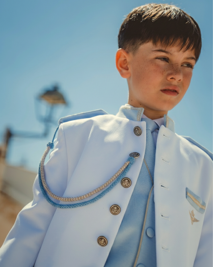 Chaqueta de comunión azul celeste de inspiración militar con cordón en el hombro y botones metálicos, presentada junto a una fachada exterior.