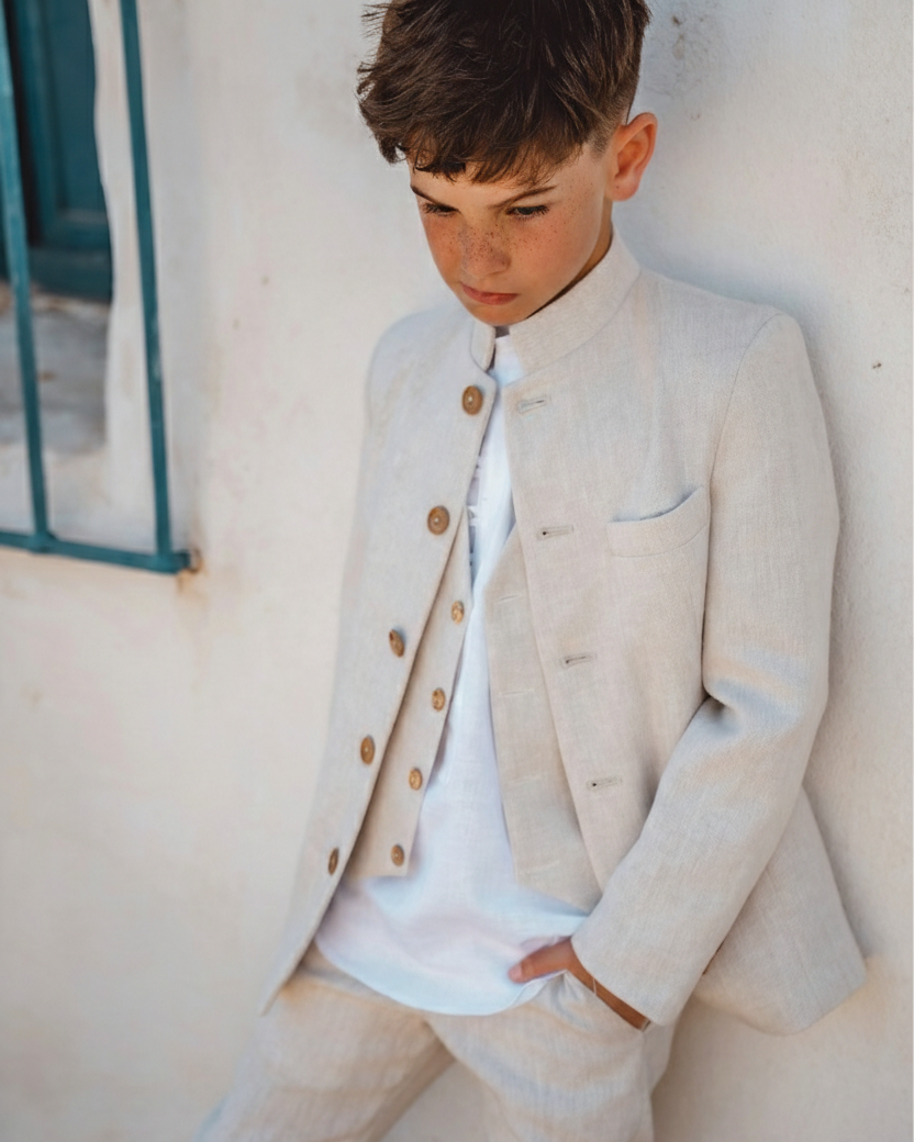 Chaqueta beige arena de comunión con cuello mao cerrado y botones dorados, combinada con camisa blanca y pantalón beige del mismo tono.