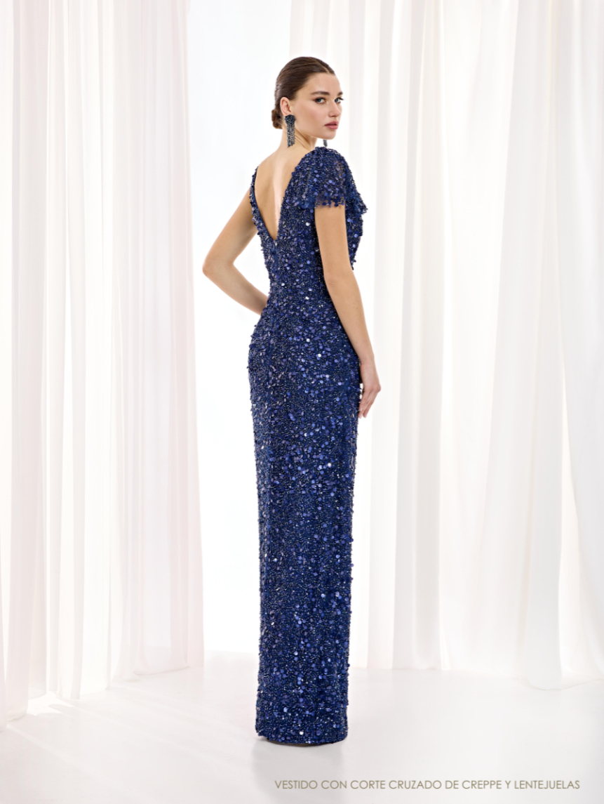 Espalda del vestido de Esthefan cubierta de lentejuelas azules, con escote en V y manga tipo capa que prolonga el brillo.
