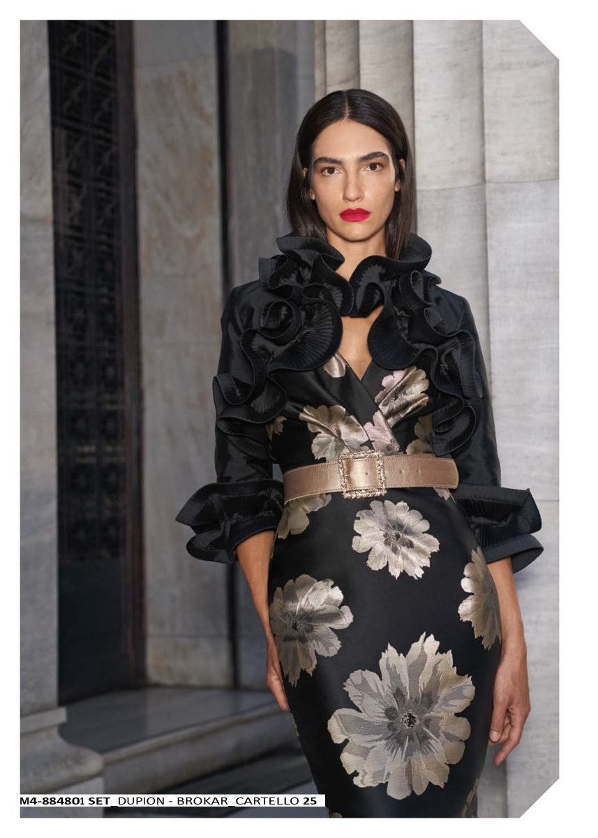 Detalle del conjunto de Mischalis con chaqueta de volantes florales negros, escote del vestido estampado y cinturón tostado de hebilla ornamentada.