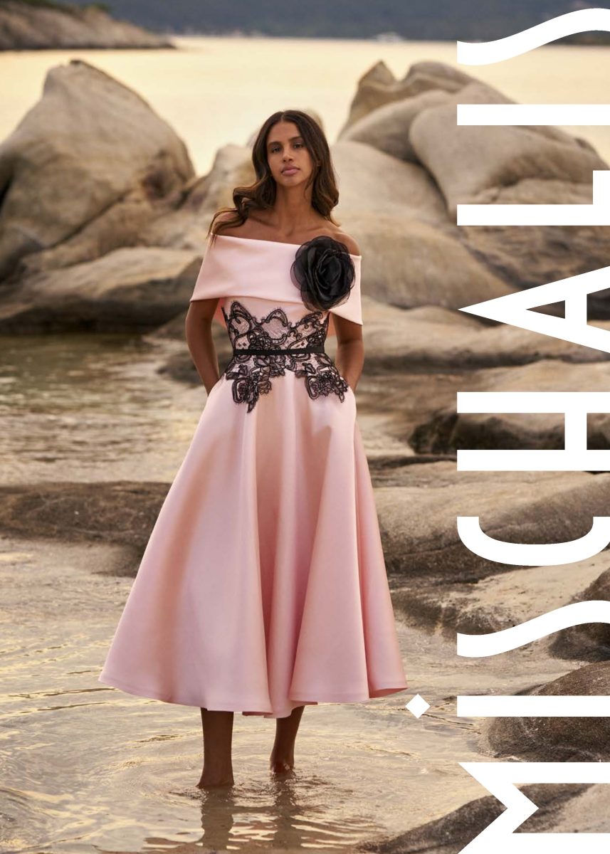 Vestido midi rosa palo de Mischalis con flor negra en el hombro, franja de encaje negro en la cintura y falda amplia con bolsillos.