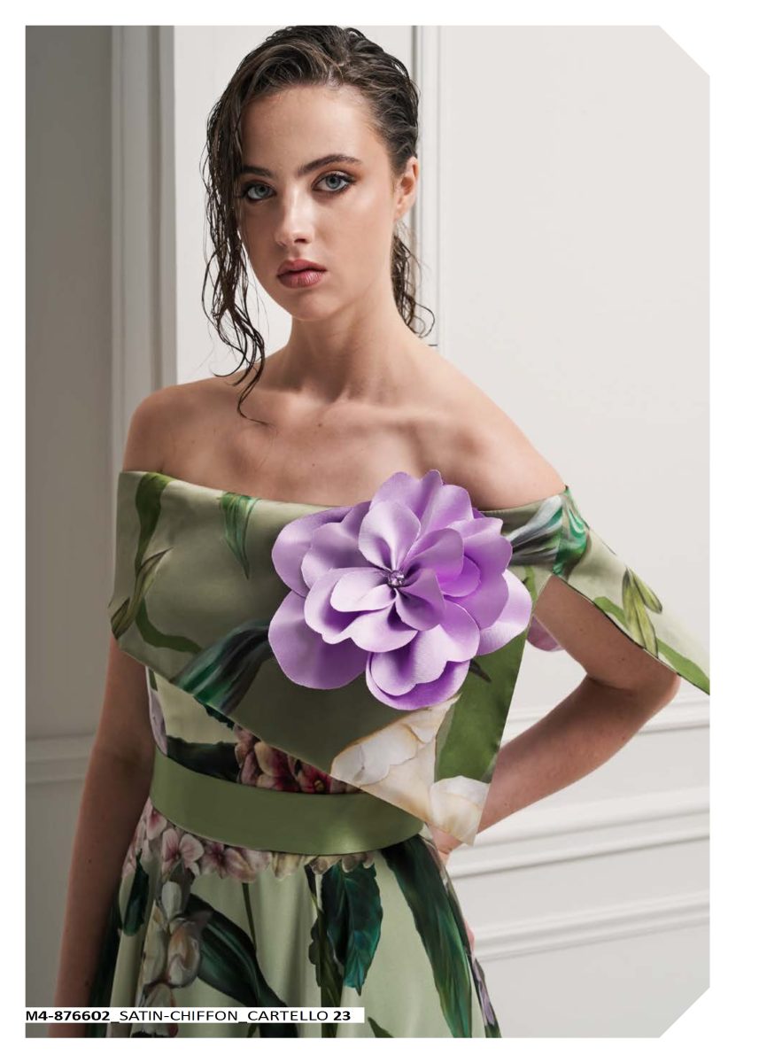 Detalle del vestido estampado de Mischalis con hombros descubiertos, capelina y gran flor lila junto al escote sobre fondo verde claro.